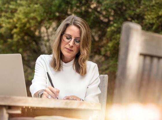 Kobieta w okularach i włosach do ramion siedzi przy stole w ogrodzie i pisze o krwawieniu po menopauzie.