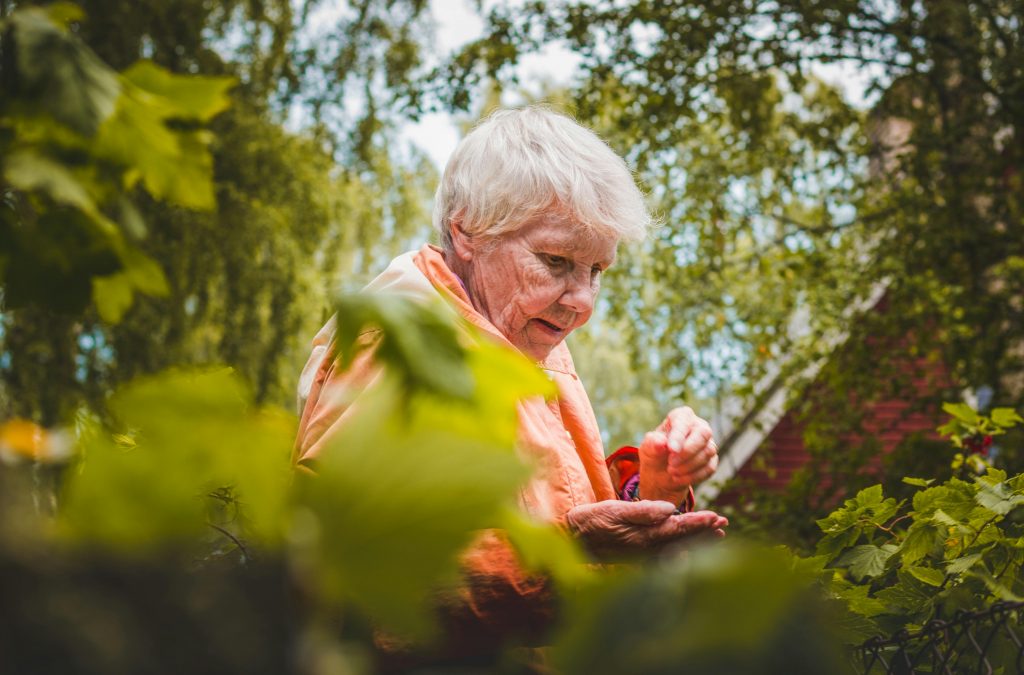 Starsza kobieta chora na chorobę Parkinsona w pomarańczowej kurtce stoi wśród zielonych liści.